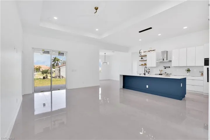 Kitchen with a raised ceiling, wall chimney exhaust hood, a kitchen island with sink, white cabinetry, and light tile patterned floors