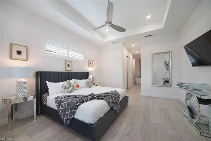 Bedroom featuring light wood finished floors, a raised ceiling, a ceiling fan, recessed lighting, and ensuite bath