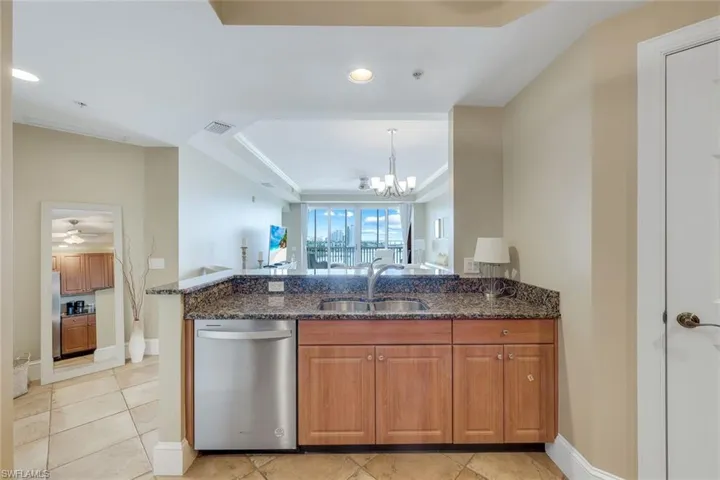 Kitchen with stainless steel dishwasher, a sink, dark stone countertops, brown cabinetry, and a raised ceiling
