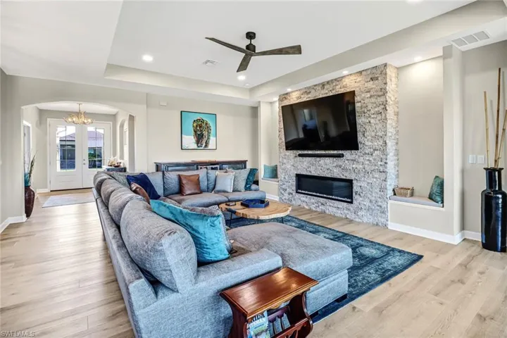 Living room featuring a stone fireplace, light wood-type flooring, ceiling fan with notable chandelier, and a raised ceiling