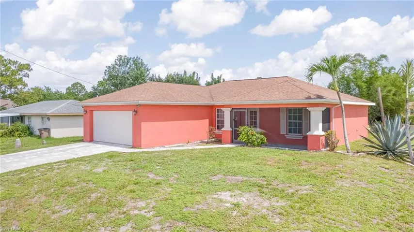 Coral-colored single-story residence featuring an attached garage with a white door, a concrete driveway, a covered entry with columns, and a front lawn with mature landscaping
