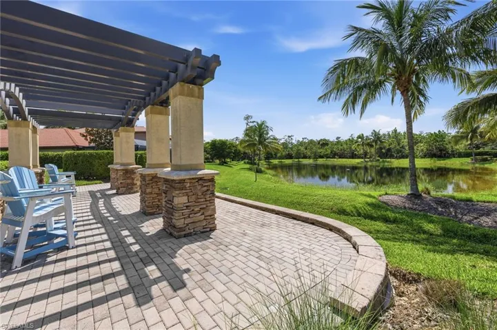 Community park paved patio with pergola, overlooking a tranquil pond, surrounded by lush green landscaping and palm trees