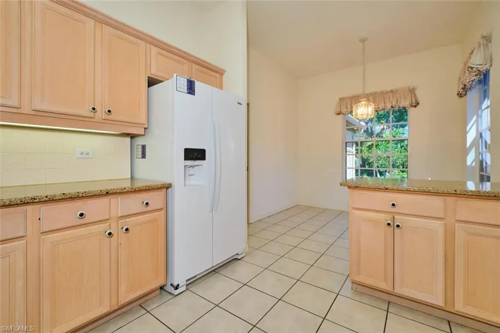 Kitchen with light wood finish cabinetry, light stone countertops, white fridge with ice dispenser, light tile patterned floors, and backsplash