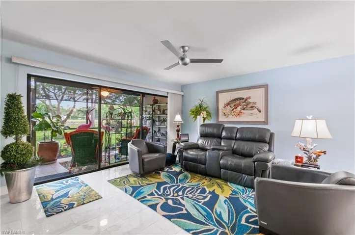 Living area featuring expansive glass sliding doors opening to an outdoor patio with lush greenery, light blue wall paint, a ceiling fan, and polished tile flooring