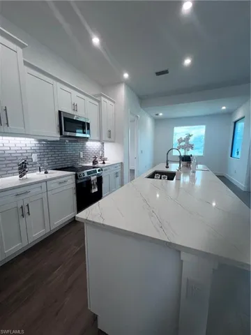 Contemporary kitchen featuring an oversized island with a built-in sink and dark pull-down faucet, white shaker cabinetry, stainless steel appliances, and wood-finish flooring