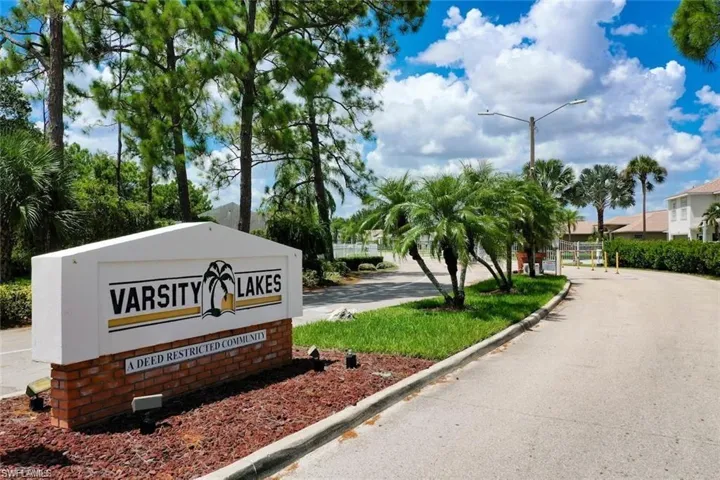 Varsity Lakes community entrance featuring a masonry monument sign, landscaped grounds, mature pine and palm trees, and paved roadway access