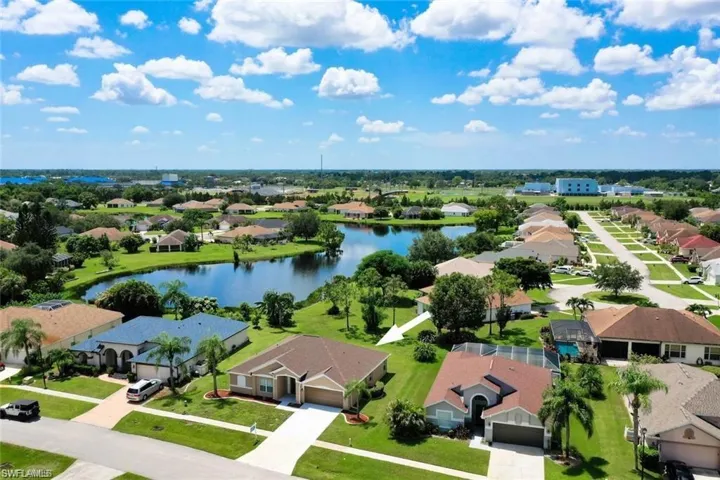 Aerial perspective featuring a single-story residence with a multi-gabled roofline, integrated two-car garage, concrete driveway, and expansive lawn area situated near a community lake