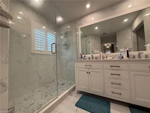 Full bath featuring light marble finish flooring, a marble finish shower, double vanity, tile walls, and recessed lighting