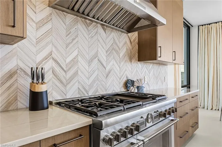 Kitchen featuring custom exhaust hood, range with two ovens, light stone countertops, modern cabinets, and tasteful backsplash - Virtually Edited Image