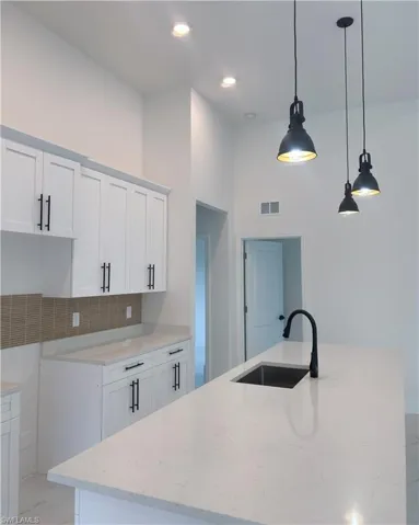 Kitchen featuring backsplash, white cabinets, decorative light fixtures, light stone countertops, and a kitchen island with sink
