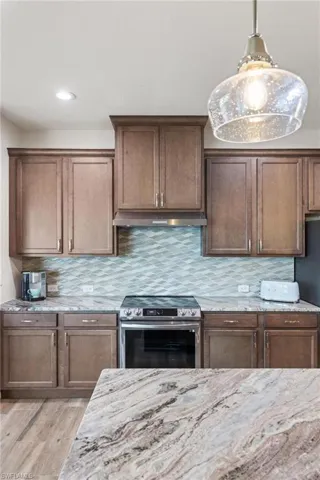 Kitchen featuring stainless steel range with electric stovetop, backsplash, hanging light fixtures, light wood-type flooring, and light stone counters