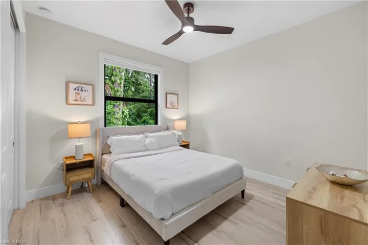 Bedroom featuring light-toned wood-finish flooring, a contemporary ceiling fan, and a window framing lush greenery