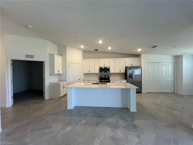 Kitchen featuring lofted ceiling, white cabinets, appliances with stainless steel finishes, a kitchen island with sink, and light stone counters