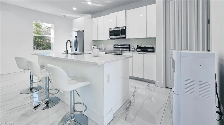 Kitchen with light marble finish flooring, modern cabinets, white cabinets, appliances with stainless steel finishes, and a kitchen island with sink