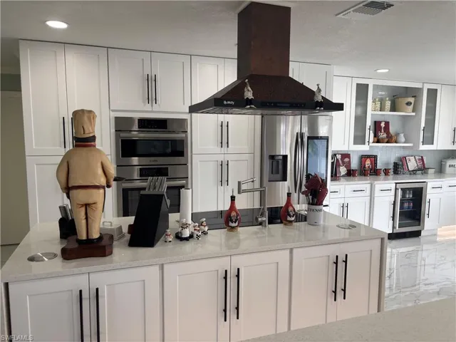 Kitchen featuring exhaust hood, beverage cooler, appliances with stainless steel finishes, white cabinetry, and glass insert cabinets
