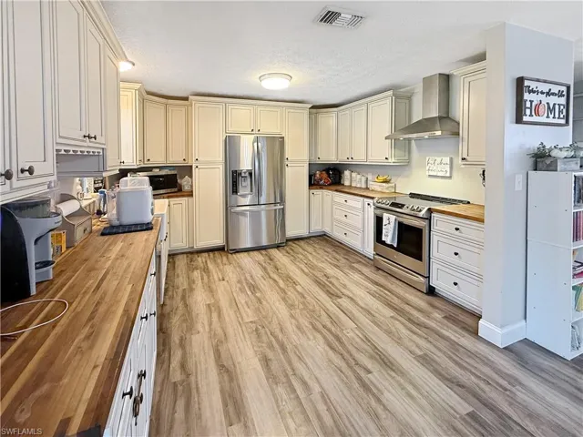 Kitchen with wood counters, wall chimney range hood, stainless steel appliances, cream cabinetry, and a textured ceiling