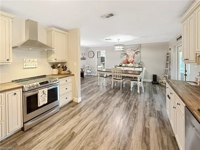 Kitchen with wooden counters, cream cabinetry, appliances with stainless steel finishes, wall chimney range hood, and a textured ceiling