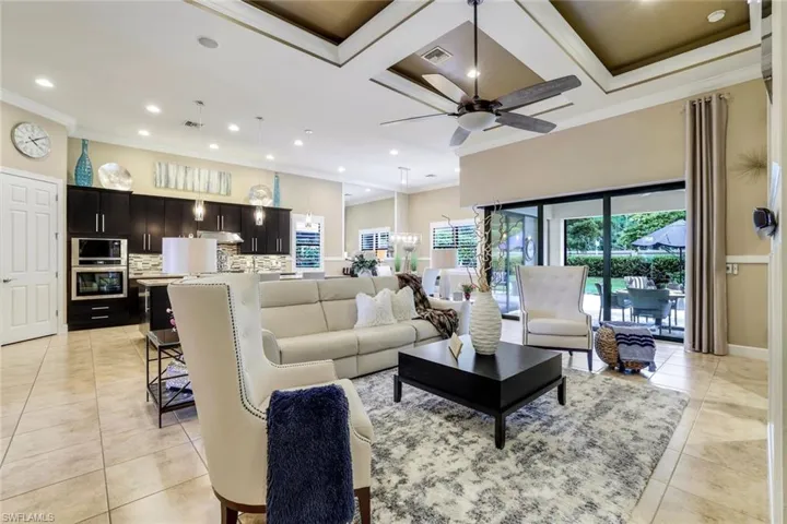 Living room featuring coffered ceiling with up to 14' ceiling, ceiling fan, crown molding, and tile flooring.  Zero corner sliding impact glass doors open the outside in for an extra expansive feel to this beautiful home.