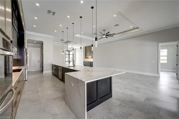 Kitchen with ornamental molding, light stone countertops, a tray ceiling, a large island with sink, and hanging light fixtures