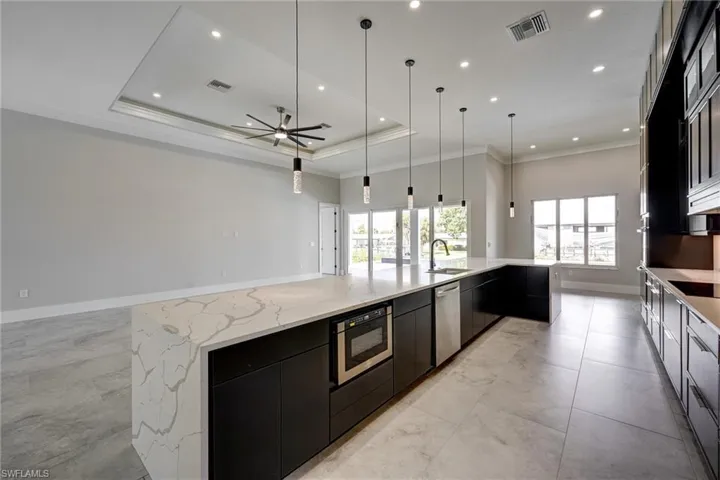 Kitchen with dark cabinets, a raised ceiling, crown molding, a large island with sink, and light stone counters
