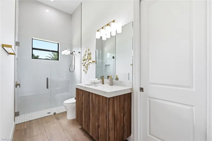 Frameless glass shower enclosure, wood-finish flooring, floating vanity with a light-toned countertop, modern vanity lighting, and a window