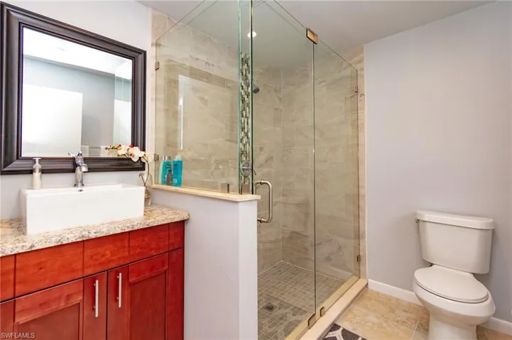 Contemporary bathroom featuring a glass-enclosed shower with tile surround and mosaic accent, a vanity with wood cabinetry and a light-toned countertop, a vessel sink, and a framed mirror