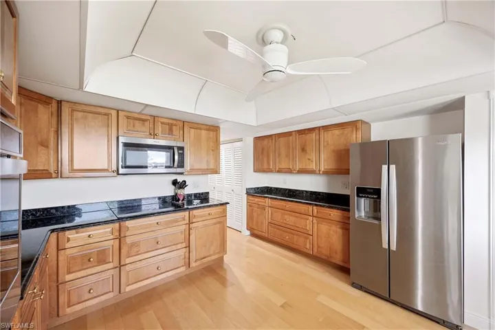Kitchen featuring appliances with stainless steel finishes, light wood finished floors, ceiling fan, dark stone counters, and brown cabinetry
