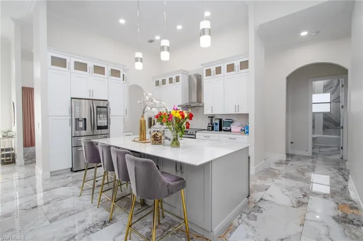 Kitchen with hanging light fixtures, wall chimney exhaust hood, an island with sink, appliances with stainless steel finishes, and white cabinetry