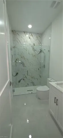 Spacious bathroom featuring a glass-enclosed shower with white and gold-veined tile, a white toilet, and a vanity with white cabinetry and a light-colored countertop