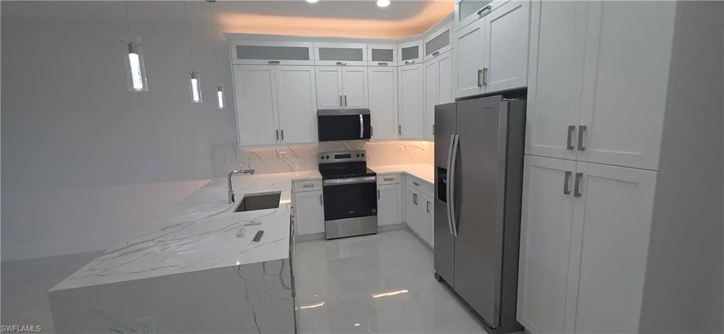 Modern kitchen featuring an expansive island with a waterfall-edge countertop and undermount sink, white shaker-style cabinetry, stainless steel appliances, and sleek pendant lighting