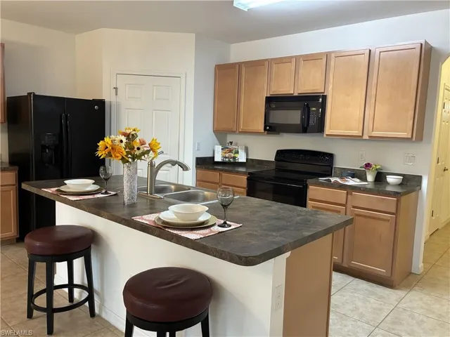 Kitchen with light tile patterned floors, dark countertops, a kitchen breakfast bar, black appliances, and a center island with sink