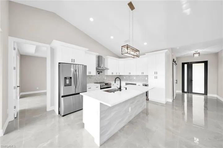 Kitchen featuring wall chimney range hood, a center island with sink, stainless steel appliances, pendant lighting, and light tile flooring