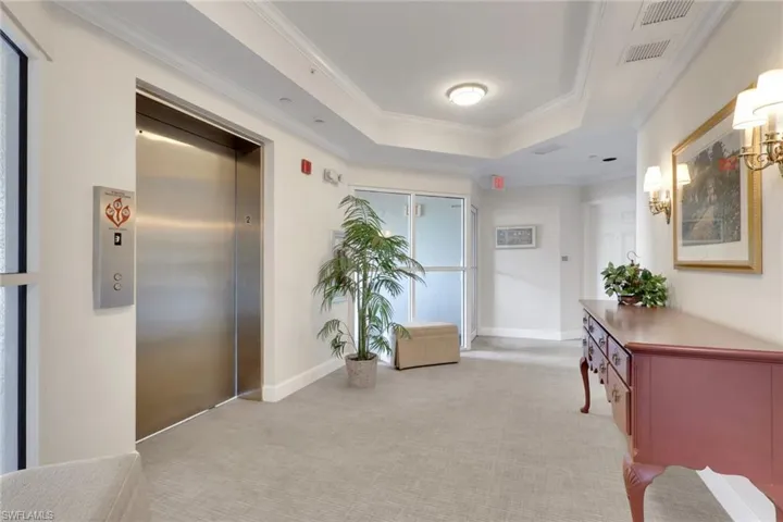 Hall featuring a raised ceiling, light colored carpet, crown molding, and elevator