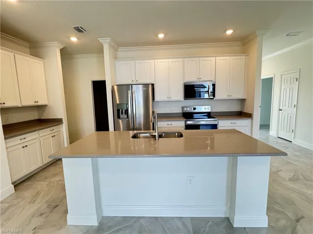 Kitchen with appliances with stainless steel finishes, a sink, light marble finish floors, an island with sink, and ornamental molding
