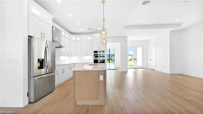 Kitchen with stainless steel appliances, wall chimney range hood, decorative light fixtures, a center island with sink, and white cabinetry