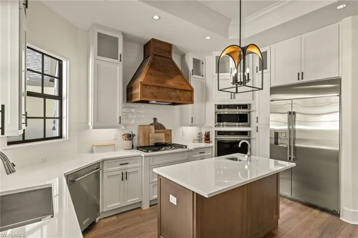 Kitchen with ornamental molding, custom range hood, stainless steel appliances, white cabinetry, and hanging light fixtures