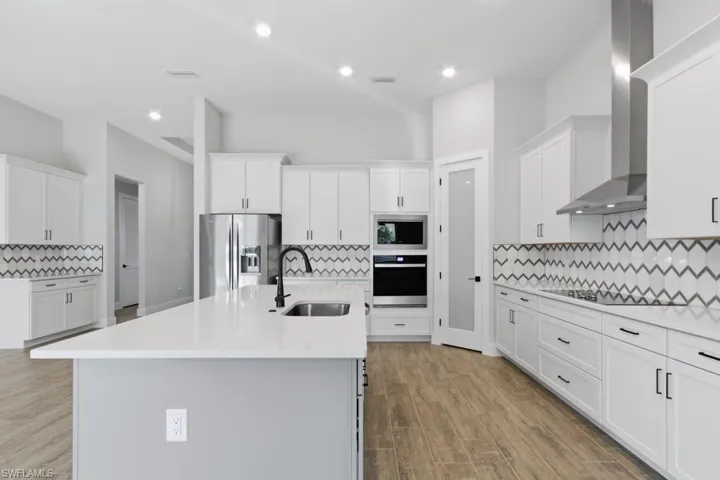 Kitchen with backsplash, white cabinetry, stainless steel appliances, wall chimney range hood, and a kitchen island with sink