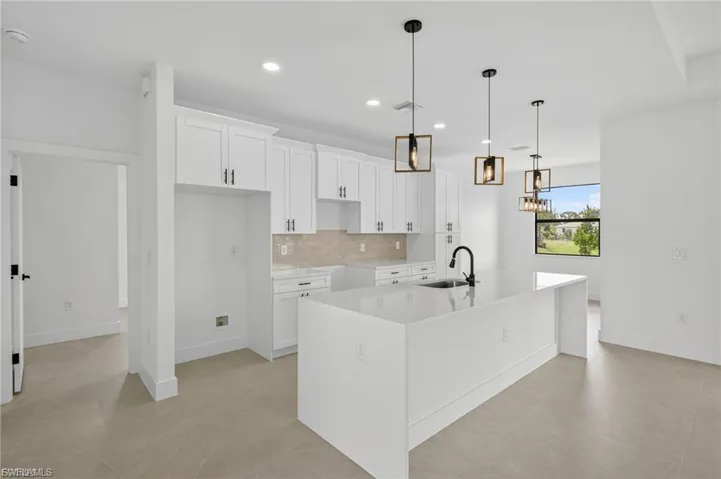 Kitchen featuring decorative light fixtures, light stone counters, an island with sink, decorative backsplash, and white cabinetry
