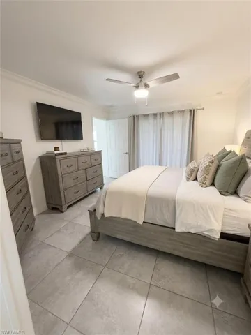 Spacious primary bedroom featuring a wood-finish bed frame, tile flooring throughout, dual dressers with ample storage, ceiling fan with integrated lighting, wall-mounted flat-screen television, and glass sliding door access to exterior space