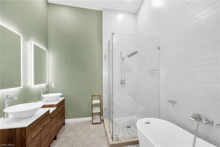 Bathroom featuring a dual vanity with vessel sinks, back-lit mirrors, a frameless glass shower enclosure with a rain shower head, a freestanding soaking tub, and hexagonal floor tiling