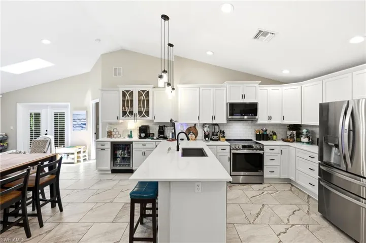 Vaulted ceiling kitchen featuring white cabinetry, stainless steel appliances, a central island with integrated sink, tile flooring, and a built-in beverage refrigerator