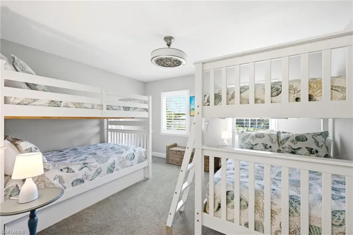 Carpeted room featuring two bunk beds with white frames, a window with white plantation shutters, and a ceiling fan with integrated lighting
