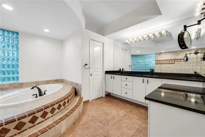 Built-in jetted tub with tile surround, glass block window, double vanity with black countertops, and a full-width wall mirror