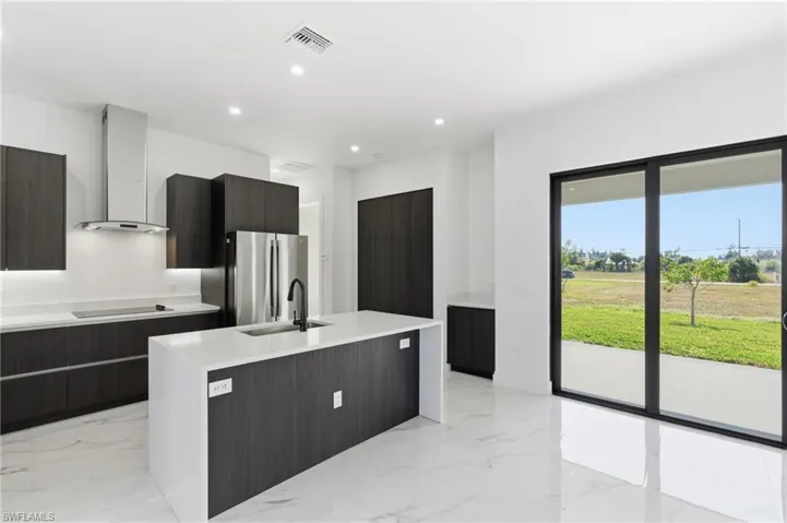 Kitchen with modern cabinets, wall chimney range hood, an island with sink, light stone counters, and freestanding refrigerator