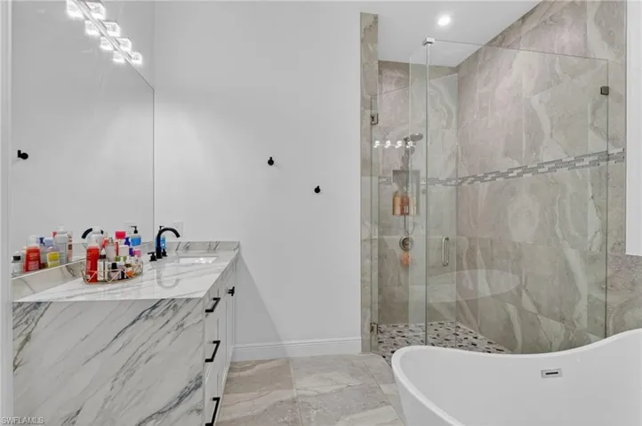 Bathroom featuring a frameless glass shower enclosure with floor-to-ceiling tile, a freestanding soaking tub, a single vanity with a stone countertop, a large mirror, and recessed lighting
