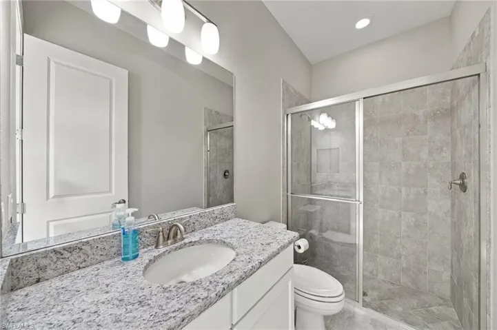 Stone-surface vanity with an integrated oval sink, full-width wall mirror, multi-bulb vanity lighting, and walk-in shower with tiled surround and metal-framed sliding glass doors