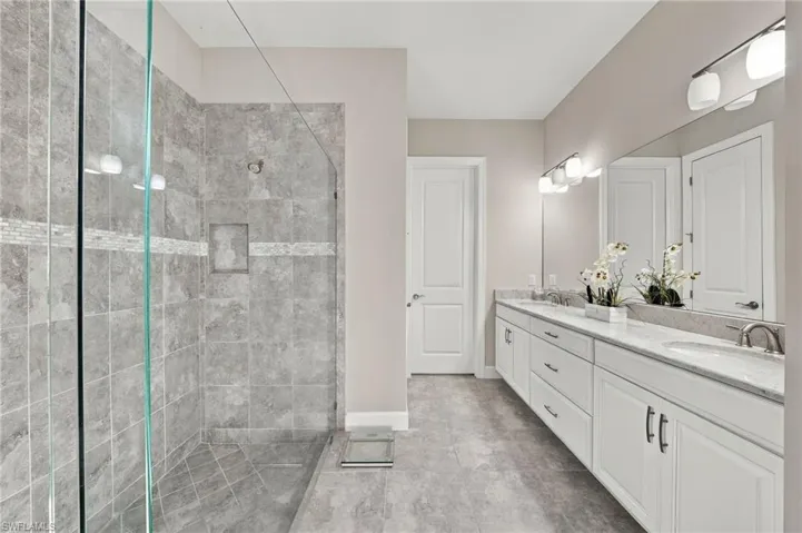 Walk-in shower with stone-finish tile surround and glass door, dual-sink vanity with stone-finish countertop, and full-width wall mirror