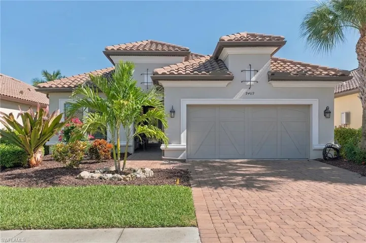 Stucco exterior with decorative crown molding and tile roof, featuring a two-car garage with carriage-style door, paved brick driveway, and landscaped front garden