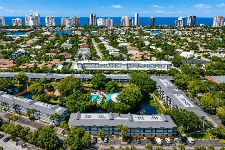 Aerial overview of a coastal community featuring multi-story residential buildings with gray roofs, surrounded by lush green landscaping and mature trees