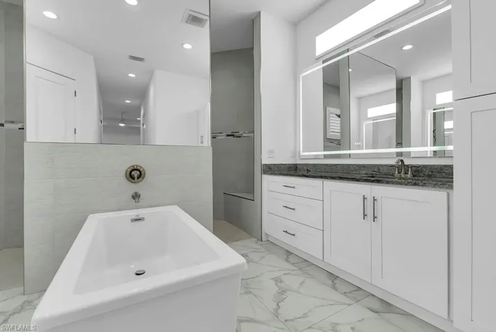 Contemporary bathroom featuring a freestanding soaking tub, large vanity with dark countertop, integrated lighted mirror, and marble-look floor tiling